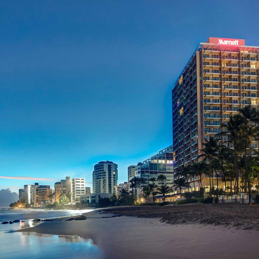 El exterior del San Juan Marriott Resort & Stellaris Casino, con la palabra "Marriott" iluminada, con la playa en primer plano y el cielo nocturno detrás.