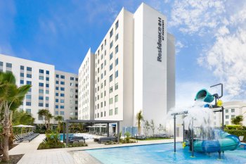 Photo showing a swimming pool with water fountain play area, with a tall white hotel building in the background. Residence Inn by Marriott San Juan Isla Verde.