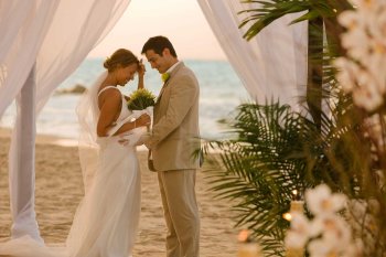 Una novia y un novio se enfrentan frente a un altar cubierto con tela blanca en una playa de Puerto Rico.