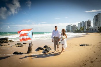 Una novia y un novio caminan por la playa cerca del San Juan Marriott Resort & Stellaris Casino, con una bandera puertorriqueña plantada en la arena.
