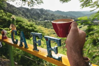 A hand holds a red mug filled with coffee in front of a mountain vista and sign that reads "Cafe."
