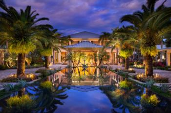 La entrada principal a un resort de lujo. Frente a la entrada hay una piscina reflectante bordeada de palmeras.
