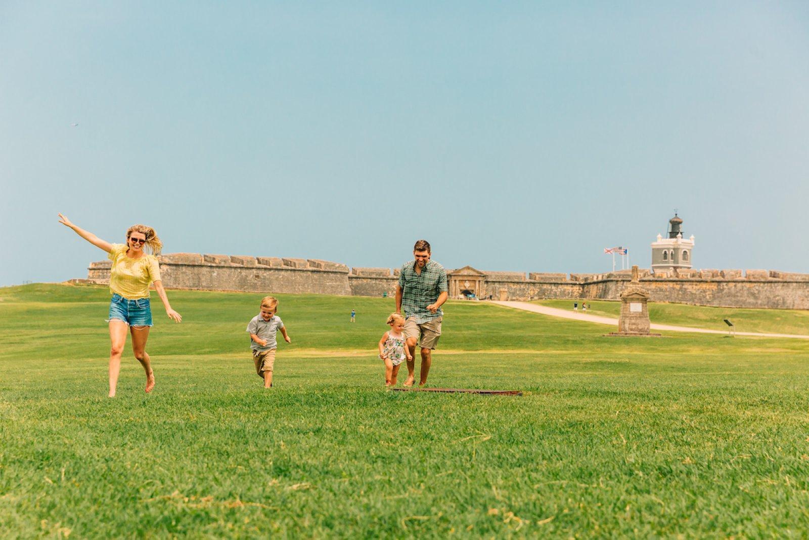 A family enjoys a sunny day at El Morro in San Juan, Puerto Rico, with children running across the green lawn.