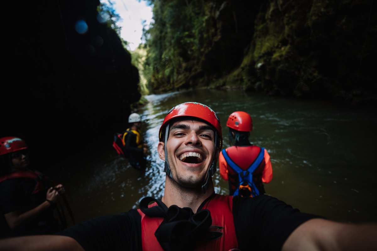 A guy in a helmet and life jacket standing in a river smiles at the camera 