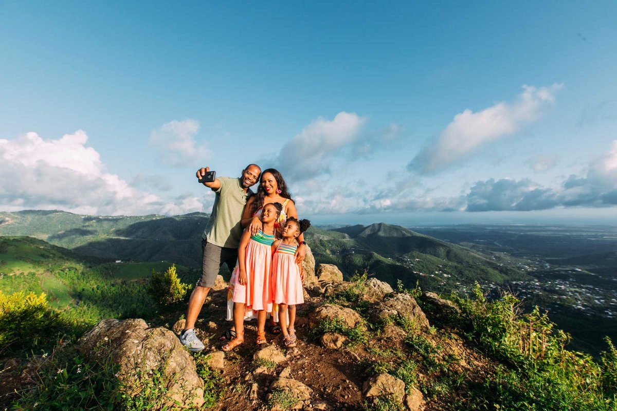 Una madre, un padre y dos niñas pequeñas posan para una selfie en la cima de una montaña con un impresionante paisaje de fondo en Puerto Rico.