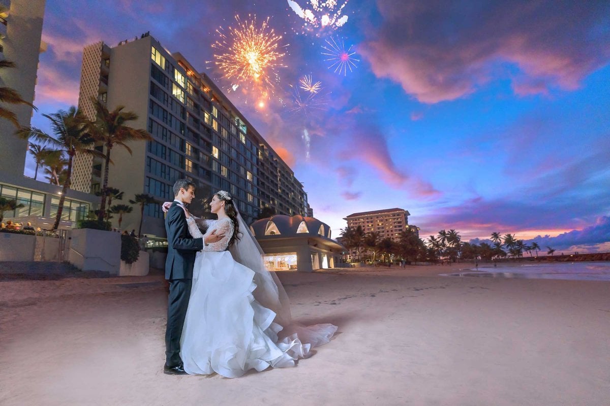 A bride and groom embrace on the beach in front of La Concha Renaissance Resort with fireworks exploding above them.