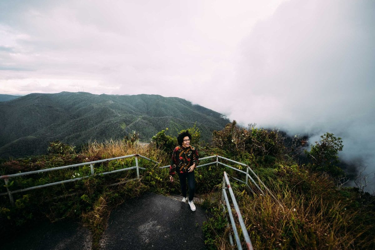 A hiker climbs a trail at Toro Negro State Forest, surrounded by low-hanging clouds and verdant mountain views.