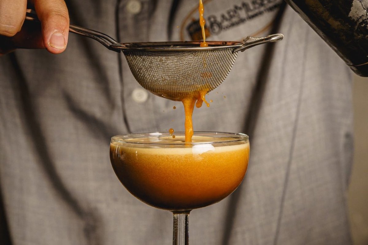 A bartender strains a cocktail into a glass. The cocktail is dark in color, like caramel or coffee. In the background is the bartender's shirt, which is embroidered with the BarMuseo logo.