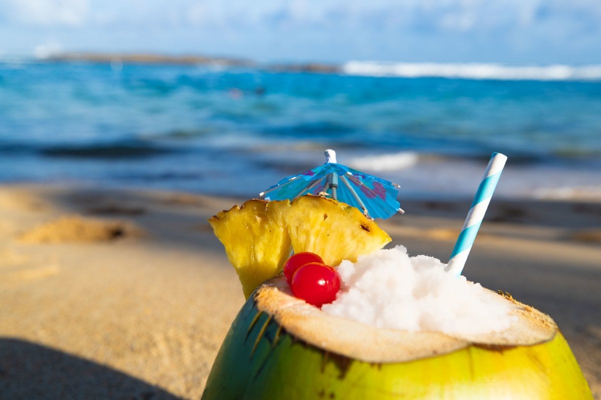 Foto de coco relleno de piña colada con una playa de fondo.