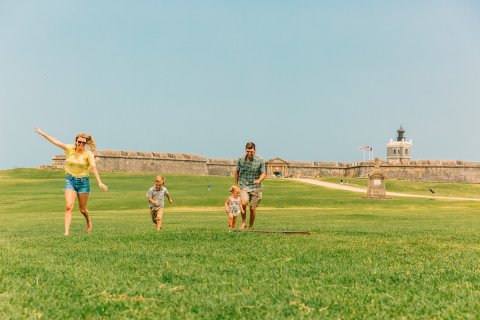 A family enjoys a sunny day at El Morro in San Juan, Puerto Rico, with children running across the green lawn.