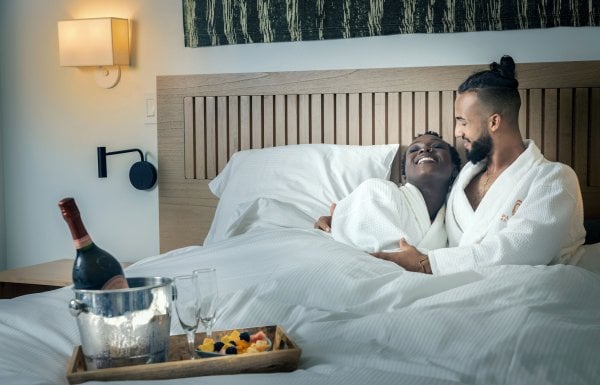 A couple lies in a hotel bed wearing white robes with a bottle of wine at the Hotel Palacio Provincial in San Juan, Puerto Rico.