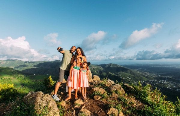 Una madre, un padre y dos niñas pequeñas posan para una selfie en la cima de una montaña con un impresionante paisaje de fondo en Puerto Rico.