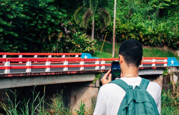 A man, seen from behind, takes a picture of a small bridge in Ciales, Puerto Rico, that is painted like the Puerto Rican flag.