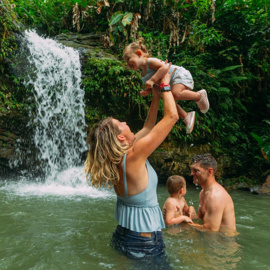 Mother holds a baby in the air while being half submerged in a forest waterfall pool.