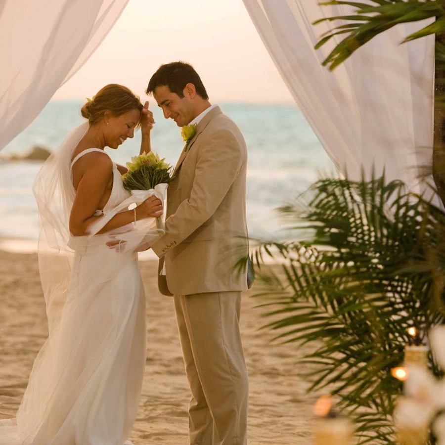 Una novia y un novio se enfrentan frente a un altar cubierto con tela blanca en una playa de Puerto Rico.