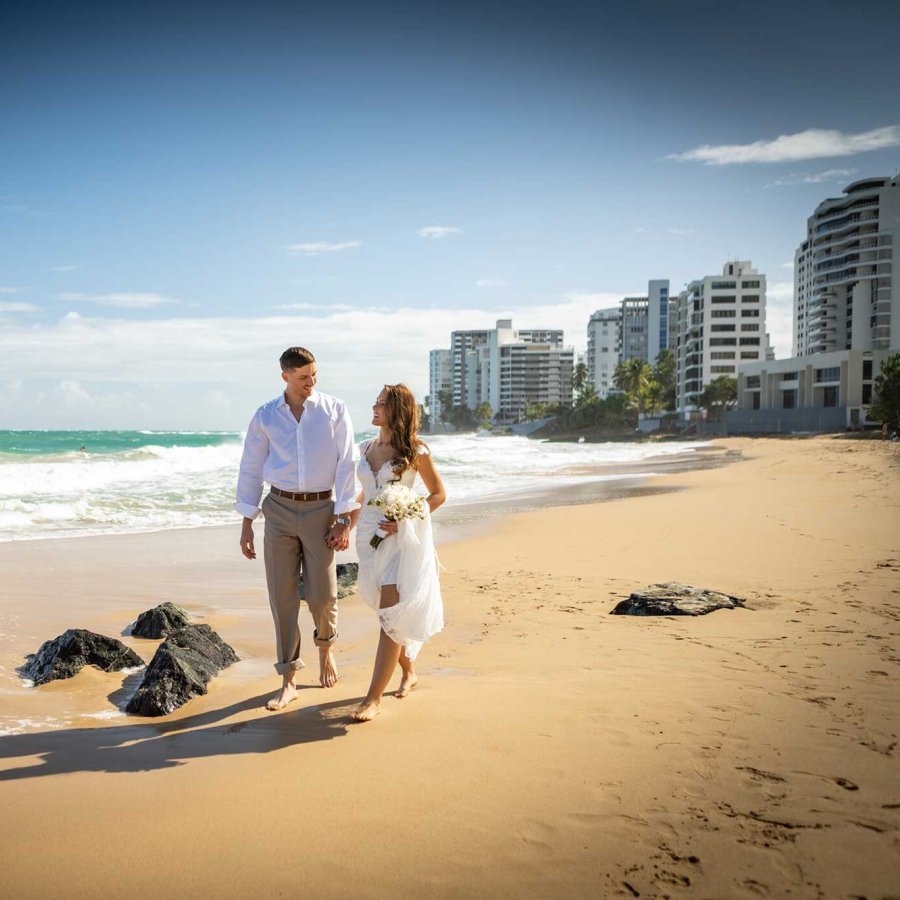 Una novia y un novio caminan por la playa cerca del San Juan Marriott Resort & Stellaris Casino, con una bandera puertorriqueña plantada en la arena.