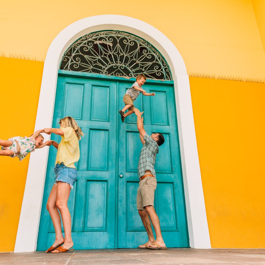 Una pareja juega con sus hijos. La familia se encuentra junto a una pared de color amarillo vivo con una puerta azul verdoso.