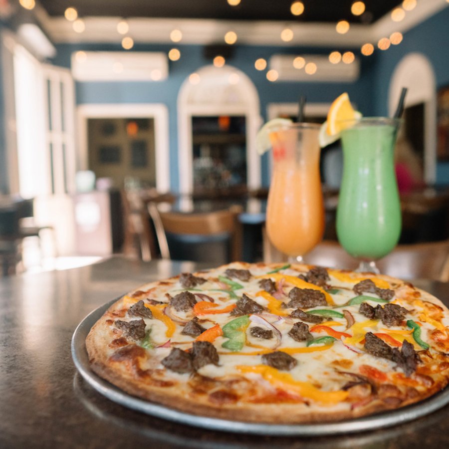 Wood-fired pizza topped with sausage and peppers alongside two frozen cocktails, one orange and the other green.