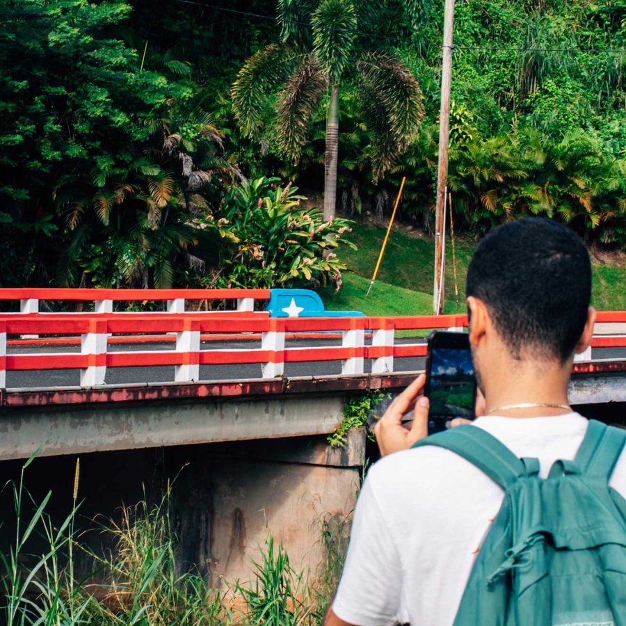 A man, seen from behind, takes a picture of a small bridge in Ciales, Puerto Rico, that is painted like the Puerto Rican flag.