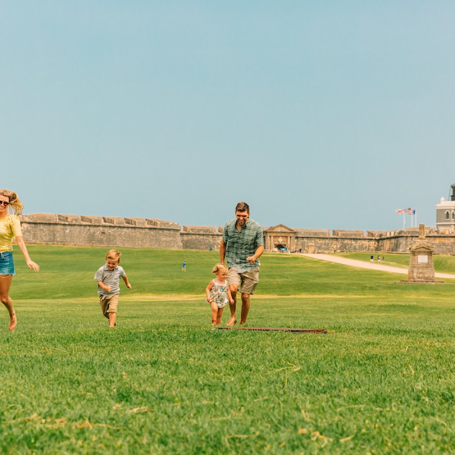 A family enjoys a sunny day at El Morro in San Juan, Puerto Rico, with children running across the green lawn.